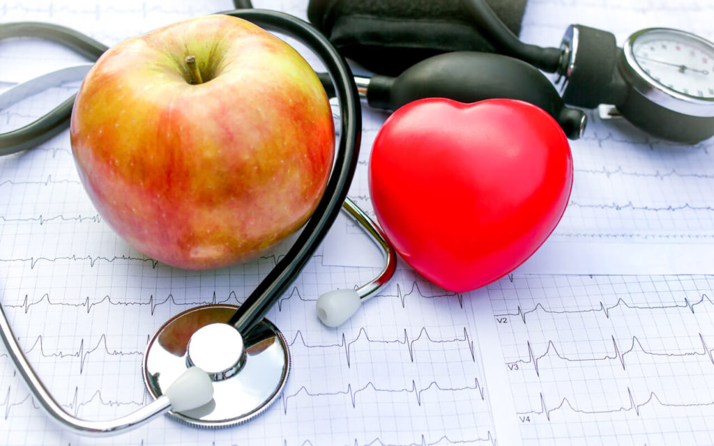a heart shaped toy, a blood pressure monitor, a stethoscope and an apple on top of a cardiogram
